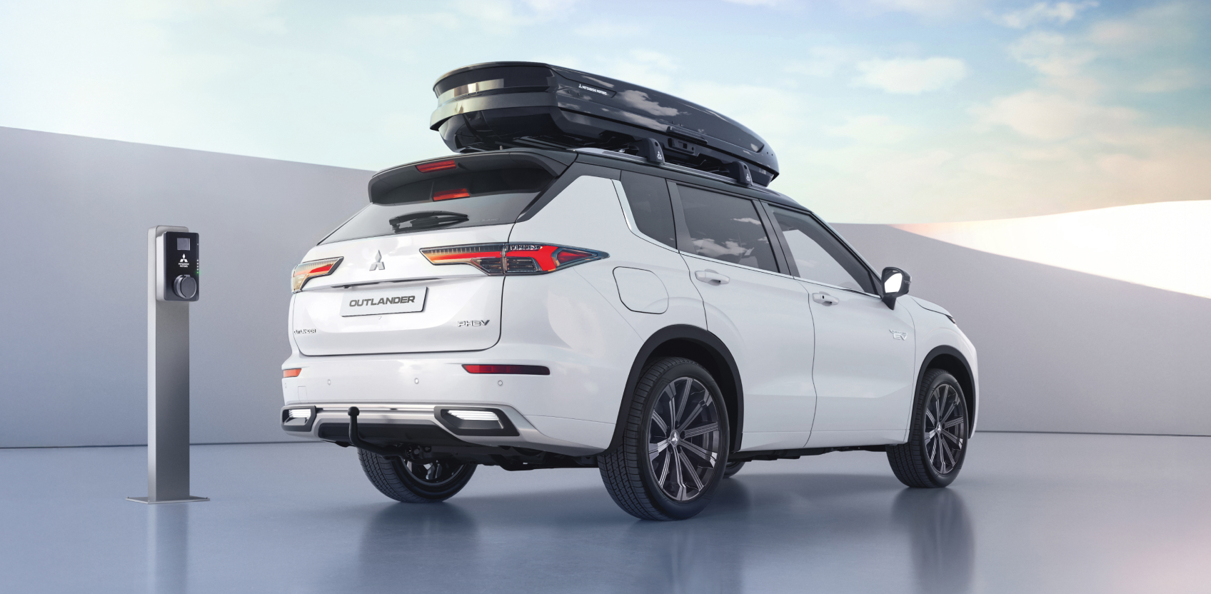 &frac34;-Heckansicht eines parkenden Outlander mit Dachgrundtr&auml;gern und Dachbox neben eine Wallbox.