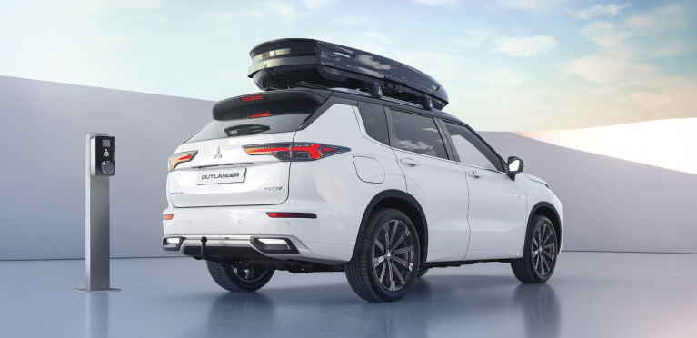 &frac34;-Heckansicht eines parkenden Outlander mit Dachgrundtr&auml;gern und Dachbox neben eine Wallbox.