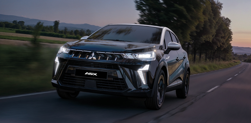 Schwarzer Mitsubishi ASX Black in dynamischer Frontansicht bei Fahrt auf einer Landstra&szlig;e bei D&auml;mmerung mit eingeschalteten LED-Scheinwerfern