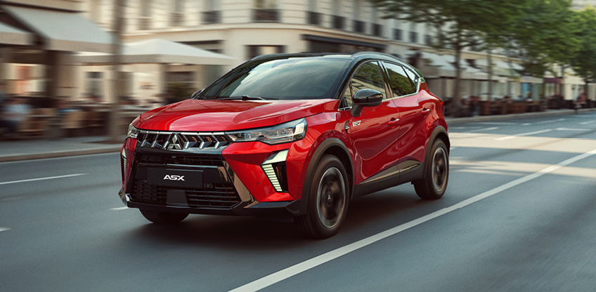 Roter Mitsubishi ASX Select in der Seitenansicht bei Fahrt durch eine Stadtstra&szlig;e mit verschwommenem Hintergrund und Geb&auml;uden