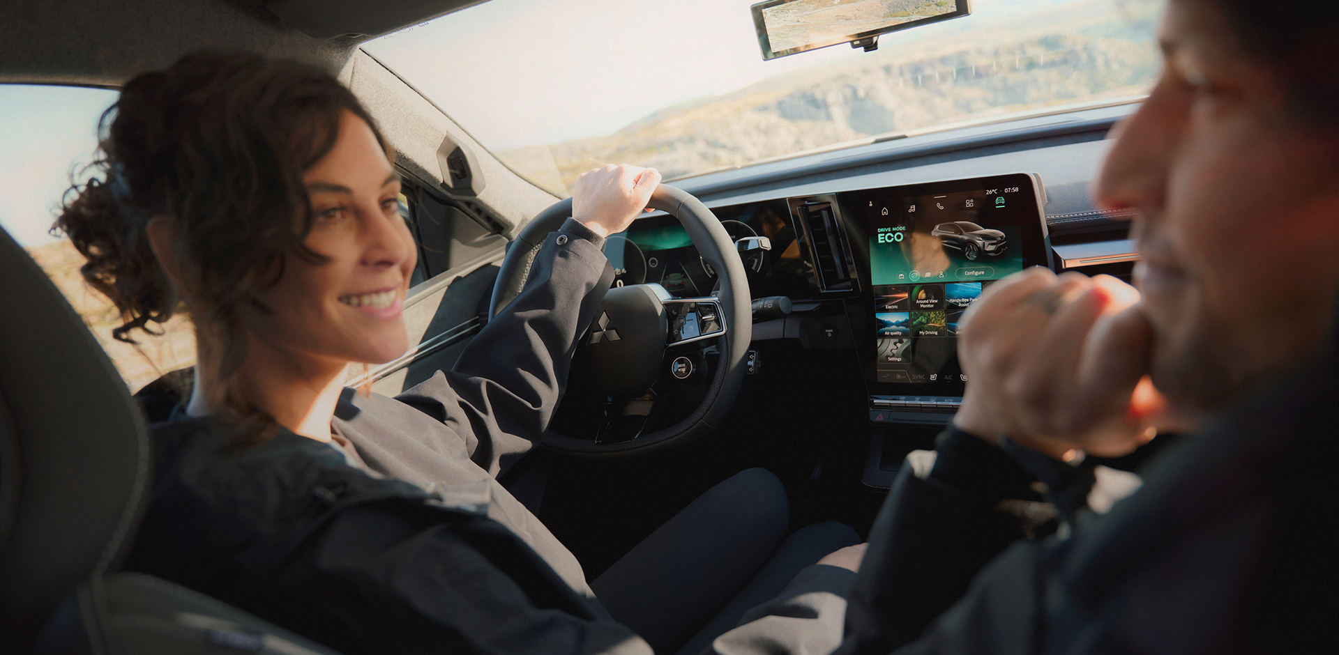 Eine Frau schaut auf dem Fahrersitz sitzend zu ihrem m&auml;nnlichen Beifahrer, auf dem Infotainment erkennt man die Fahreinstellung ECO.