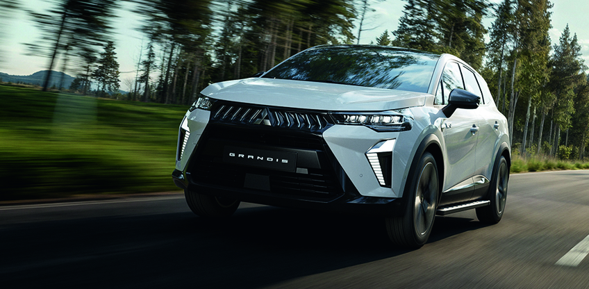 Wei&szlig;er Mitsubishi Grandis SUV f&auml;hrt dynamisch auf einer Landstra&szlig;e durch einen Wald, Frontansicht in Bewegung.