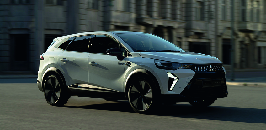Wei&szlig;er Mitsubishi Grandis SUV f&auml;hrt dynamisch durch eine Stadt, dreiviertel Frontansicht mit unscharfem Hintergrund.