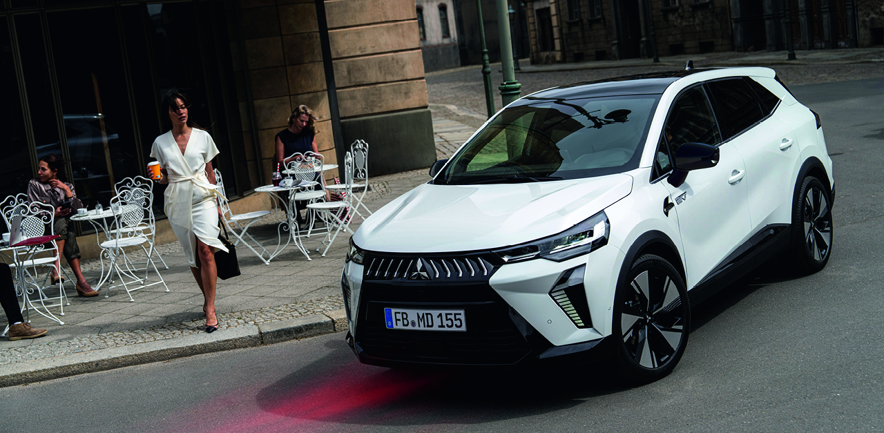Wei&szlig;er Mitsubishi Grandis SUV in der Stadt vor einem Stra&szlig;encaf&eacute;, Frau in wei&szlig;em Kleid mit Kaffeebecher geht am Fahrzeug vorbei
