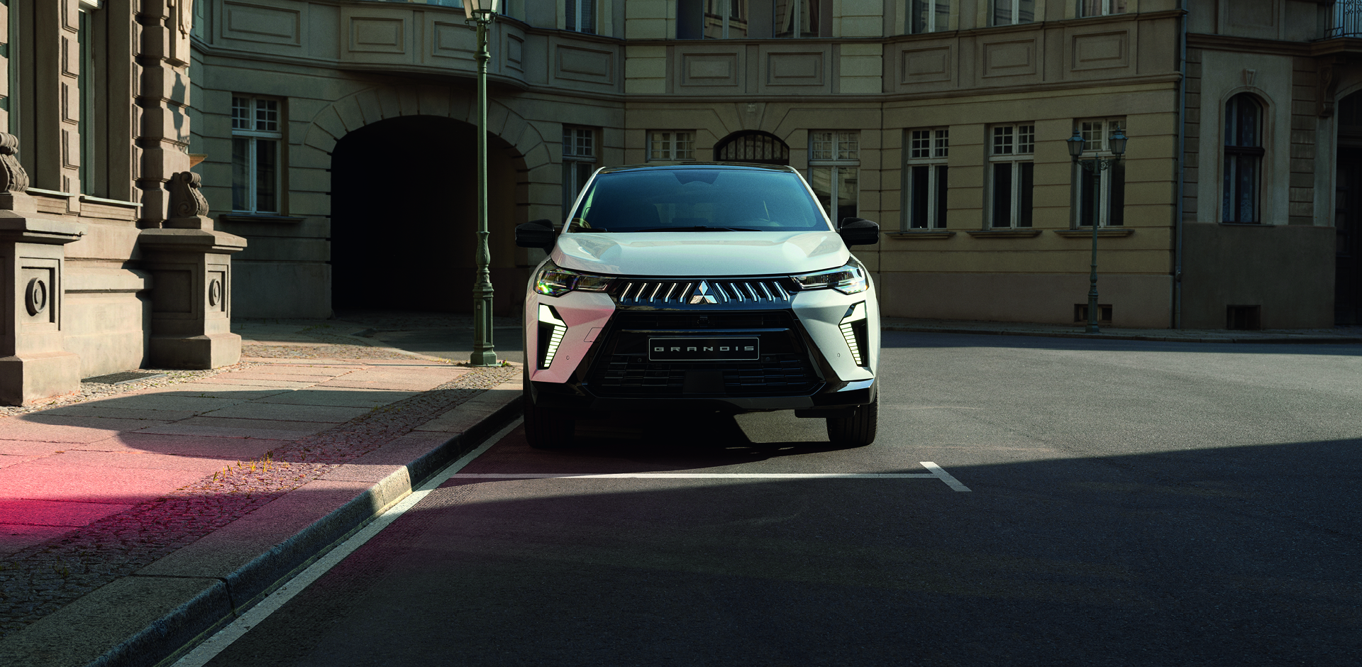 Wei&szlig;er Mitsubishi Grandis steht frontal geparkt an einer ruhigen Stadtstra&szlig;e vor einem historischen Geb&auml;ude.