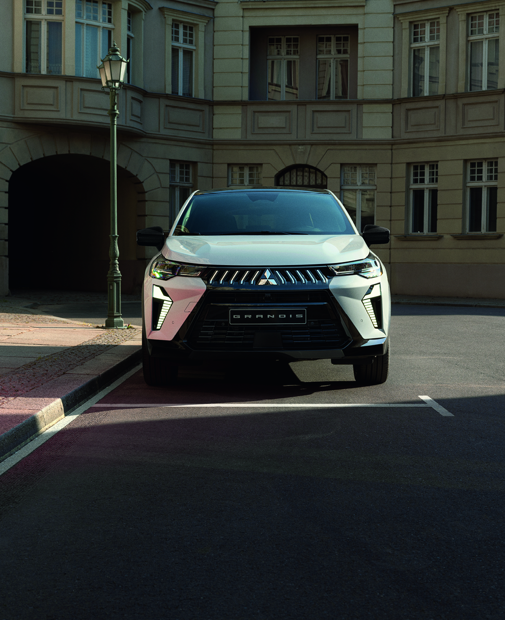 Wei&szlig;er Mitsubishi Grandis steht frontal geparkt an einer ruhigen Stadtstra&szlig;e vor einem historischen Geb&auml;ude.