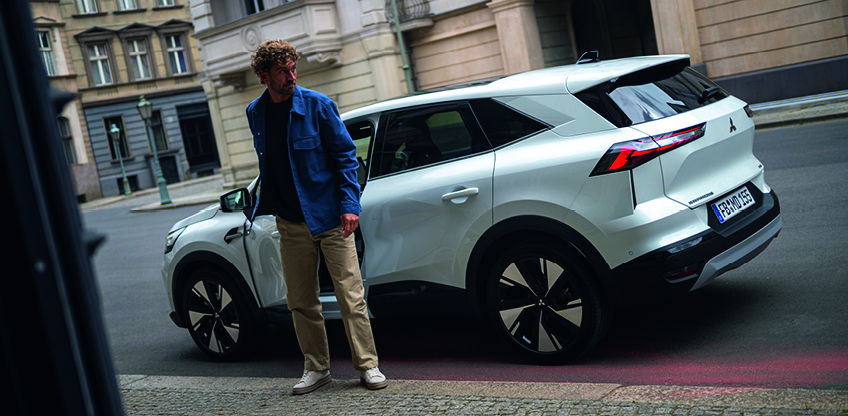 Mann steigt aus einem wei&szlig;en Mitsubishi Grandis SUV am Stra&szlig;enrand in der Stadt aus und sieht sich um, Fahrzeug in schr&auml;ger R&uuml;ckansicht geparkt