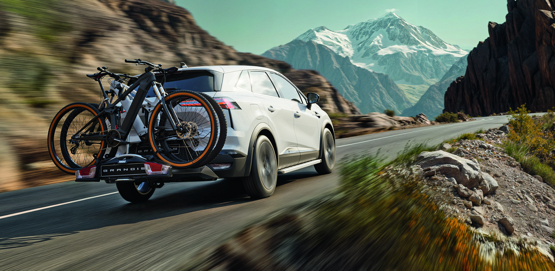 Wei&szlig;er Mitsubishi Grandis f&auml;hrt mit montiertem Heck-Fahrradtr&auml;ger und zwei Fahrr&auml;dern durch eine Berglandschaft auf einer Landstra&szlig;e.