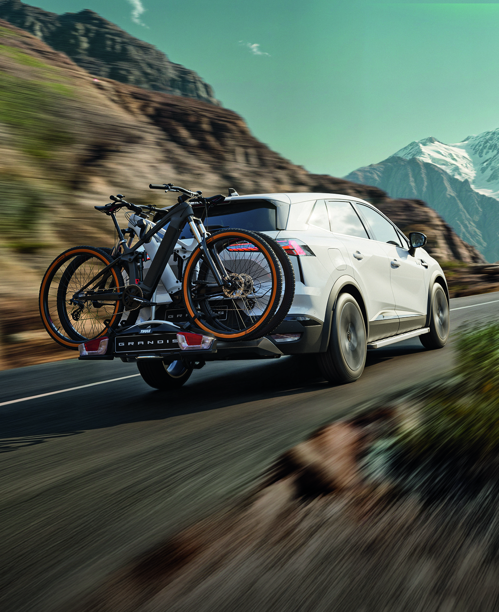 Wei&szlig;er Mitsubishi Grandis f&auml;hrt mit montiertem Heck-Fahrradtr&auml;ger und zwei Fahrr&auml;dern durch eine Berglandschaft auf einer Landstra&szlig;e.