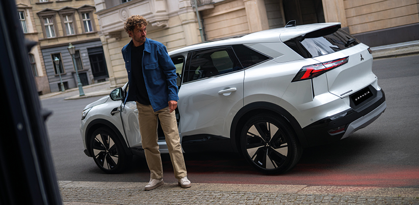 Ein Mann in Jeansjacke steht neben einem wei&szlig;en Mitsubishi Grandis Intro Edition vor einem Geb&auml;ude