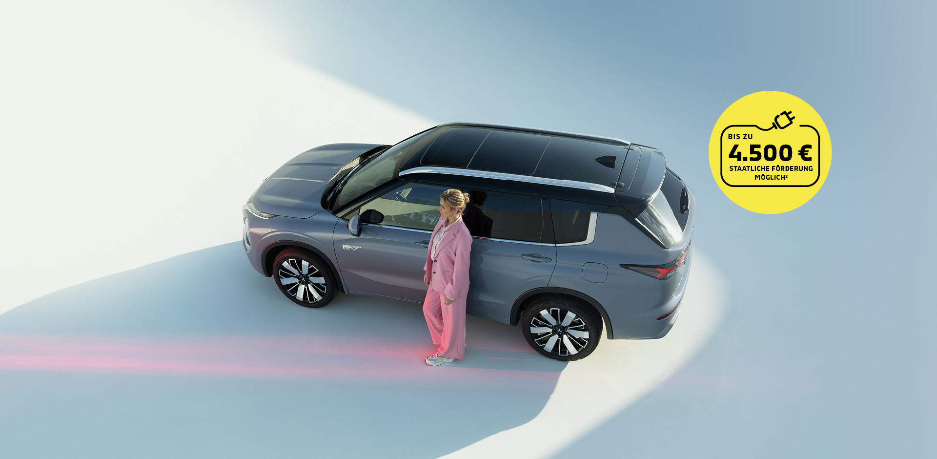Grauer Mitsubishi Outlander Plug-In Hybrid SUV von oben, Frau in rosafarbenem Anzug steht neben dem Fahrzeug. Gelber Kreis mit Hinweis auf staatliche F&ouml;rderung bis zu 4.500 Euro.