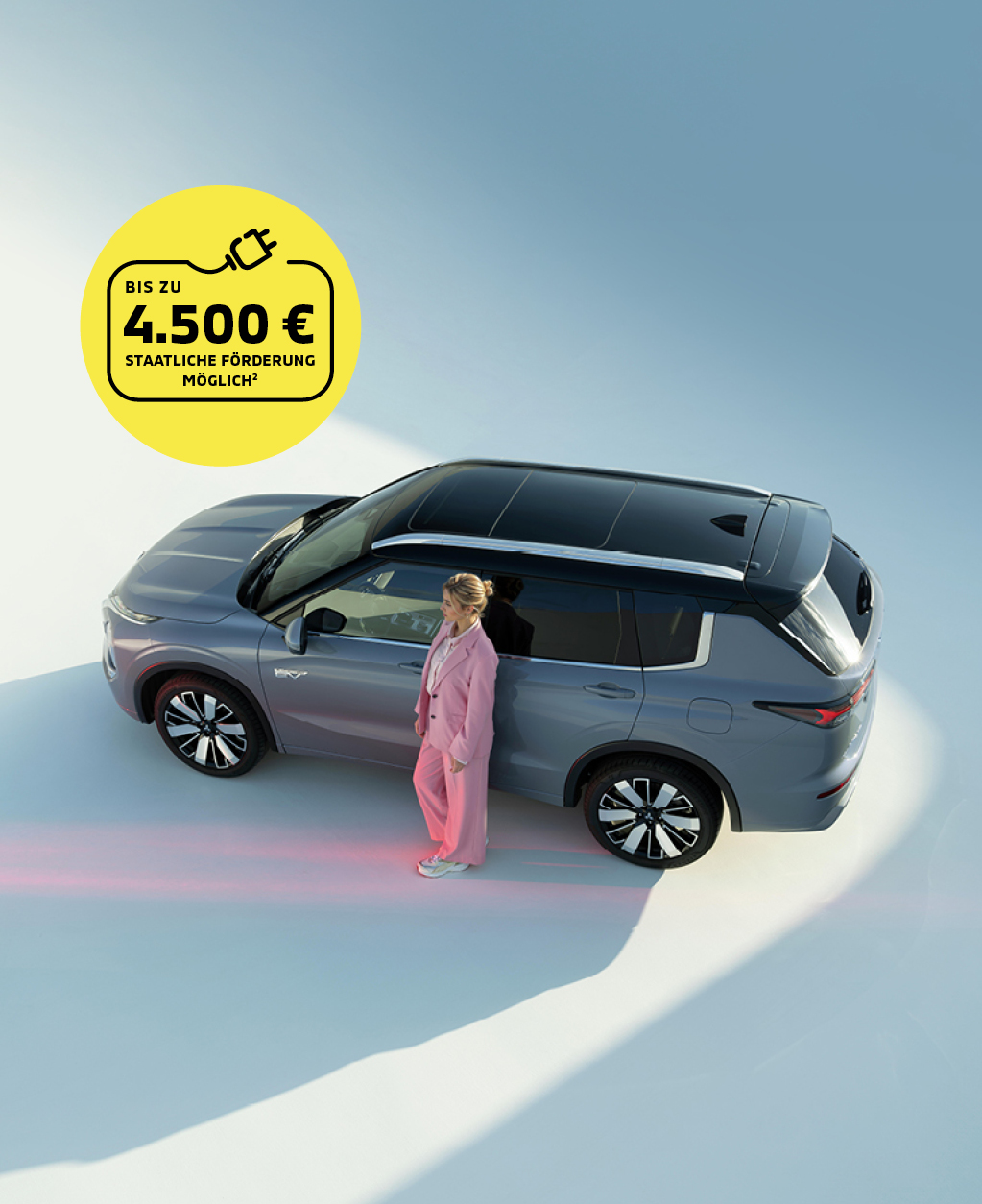 Grauer Mitsubishi Outlander Plug-In Hybrid SUV von oben, Frau in rosafarbenem Anzug steht neben dem Fahrzeug. Gelber Kreis mit Hinweis auf staatliche F&ouml;rderung bis zu 4.500 Euro.