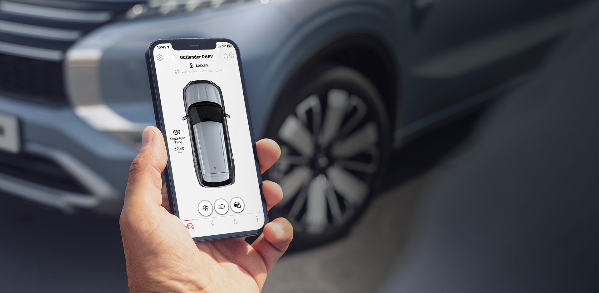 Eine Hand h&auml;lt ein Smartphone mit ge&ouml;ffneter MITSUBISHI MOTORS App, im Hintergrund ist ein Outlander zu sehen.