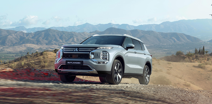 Silberner Mitsubishi Outlander Select in der Frontansicht auf unbefestigtem Gel&auml;nde mit Bergpanorama im Hintergrund