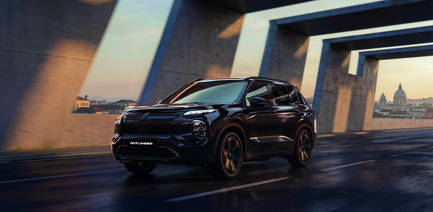 Schwarzer Mitsubishi Outlander Black in dynamischer Frontansicht unter einem modernen Br&uuml;ckenbauwerk im urbanen Stadtumfeld bei Abendd&auml;mmerung