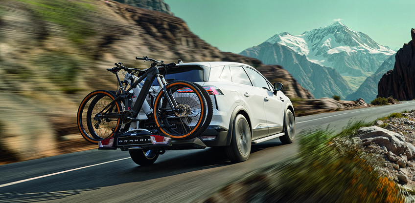Wei&szlig;er Mitsubishi Grandis mit montiertem Fahrradhecktr&auml;ger und zwei Fahrr&auml;dern f&auml;hrt dynamisch auf einer Bergstra&szlig;e, im Hintergrund schneebedeckte Gipfel.