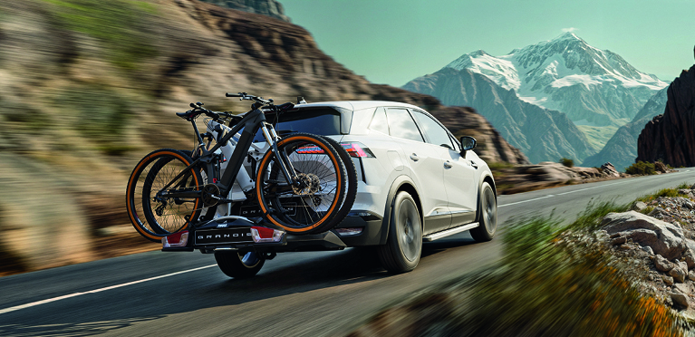 Wei&szlig;er Mitsubishi Grandis mit montiertem Fahrradhecktr&auml;ger und zwei Fahrr&auml;dern f&auml;hrt dynamisch auf einer Bergstra&szlig;e, im Hintergrund schneebedeckte Gipfel.