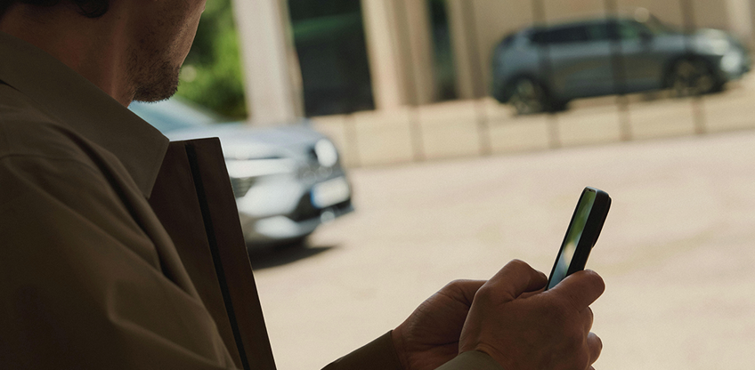 Im Vordergrund h&auml;lt ein Mann ein Mobiltelefon in der Hand, w&auml;hrend sich der Eclipse Cross im Hintergrund in der Glasfront eines modernen Geb&auml;udes spiegelt.