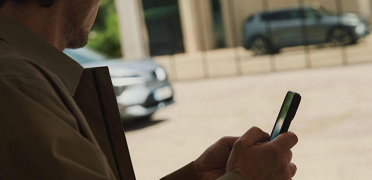 Im Vordergrund h&auml;lt ein Mann ein Mobiltelefon in der Hand, w&auml;hrend sich der Eclipse Cross im Hintergrund in der Glasfront eines modernen Geb&auml;udes spiegelt.